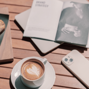 Flat lay of a coffee on a wooden table with an open brand strategy book and phone, styled in warm Auburn tones to represent brand confidence and clarity.