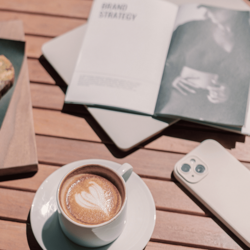 Flat lay of a coffee on a wooden table with an open brand strategy book and phone, styled in warm Auburn tones to represent brand confidence and clarity.